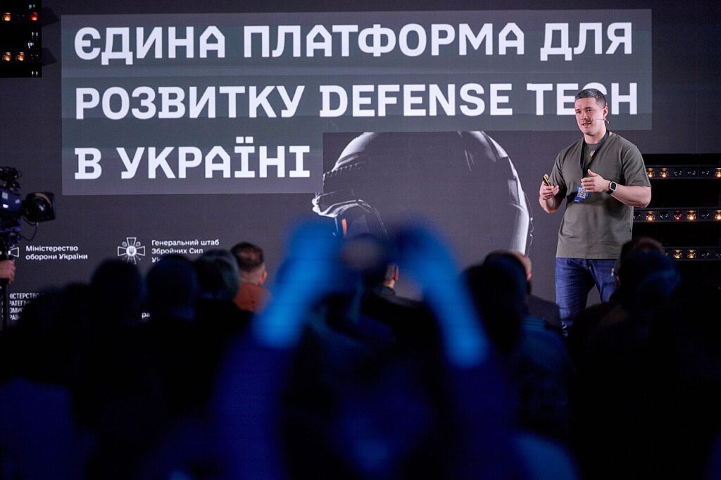 A speaker addresses an audience, seen from behind, in front of a screen reading “A single platform for Ukraine's defense-tech development”