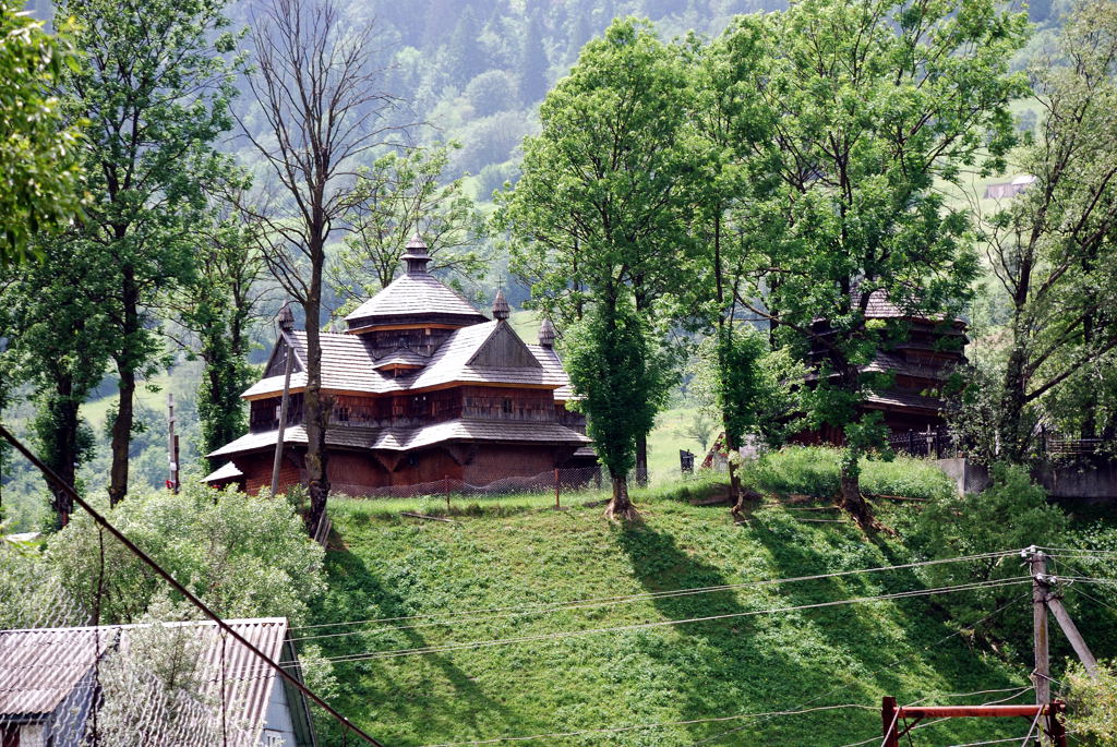 A cross-shaped wooden church with a tall central roof, standing on a hill surrounded by trees