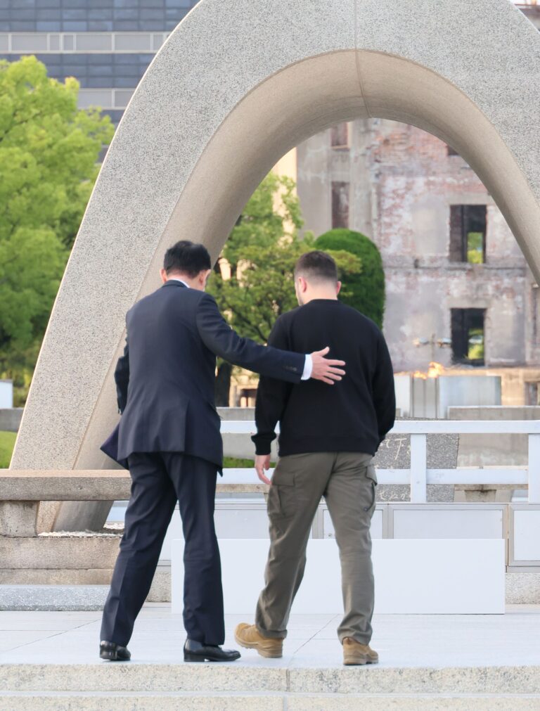 原爆死没者慰霊碑のアーチの下を歩くゼレンスキー大統領の背中に、岸田総理が手を添えている。アーチの先に原爆ドームが見える