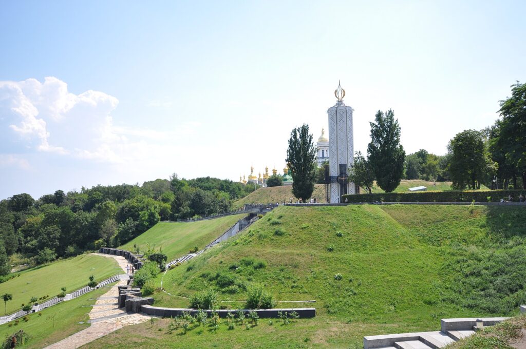 A slender monument, the “Candle of Memory,” standing on a hill. In front, a row of gravestone-like memorial stones honoring the victims; in the background, the golden domes of the Kyiv Pechersk Lavra