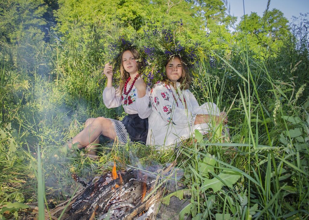 Two girls wearing embroidered shirts and flower wreaths on their heads, sitting beside a bonfire