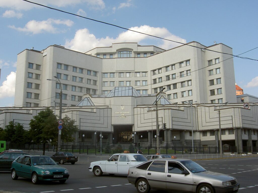 The front of Ukraine's Constitutional Court building, marked “КОНСТИТУЦІЙНИЙ СУД УКРАЇНИ,” photographed across a street with passing traffic